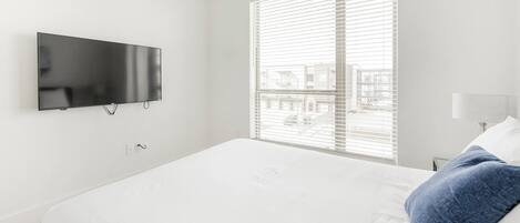 A modern hotel room with a large bed, a flat-screen TV mounted on the wall, and a window with blinds.