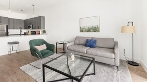 Uma sala de estar moderna com um sofá cinzento, uma poltrona verde, uma mesa de centro de vidro e um abajur.