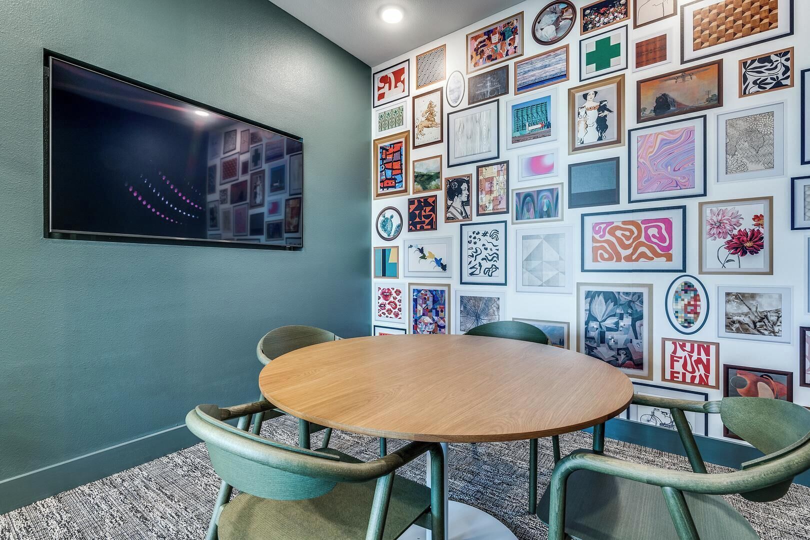 A conference room with a round table and four chairs, a flat-screen TV mounted on the wall, and a wall adorned with various framed artworks.