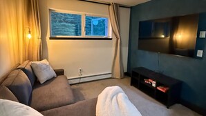 A modern living room with a grey sofa, a white throw blanket, a flat-screen TV mounted on the wall, and a window with curtains.