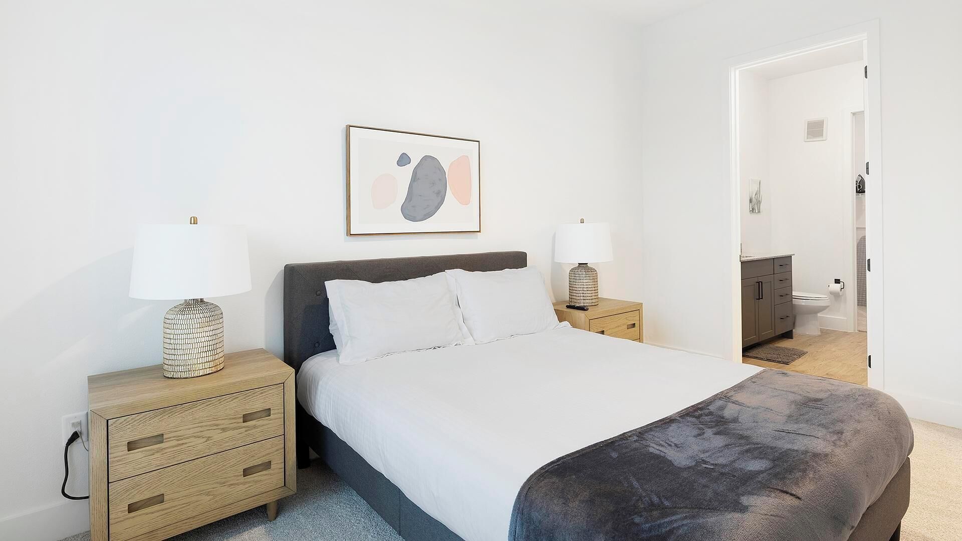 A bedroom with a bed, two bedside tables, a nightstand, a framed picture on the wall, and a bathroom visible through an open door.