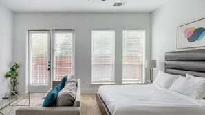 A modern bedroom with a large bed, a seating area with a sofa and cushions, and three windows with blinds. - Landing | Beautiful Studio, Courtyard, Clubhouse (Denton)