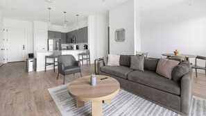 A modern living room with a grey sofa, a round wooden coffee table, and a kitchen in the background. - Landing | Amazing 3BD, Pool, Courtyard (Denton)