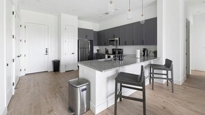 A modern kitchen with a central island, stainless steel appliances, and a trash can. - Landing | Amazing 3BD, Pool, Courtyard (Denton)