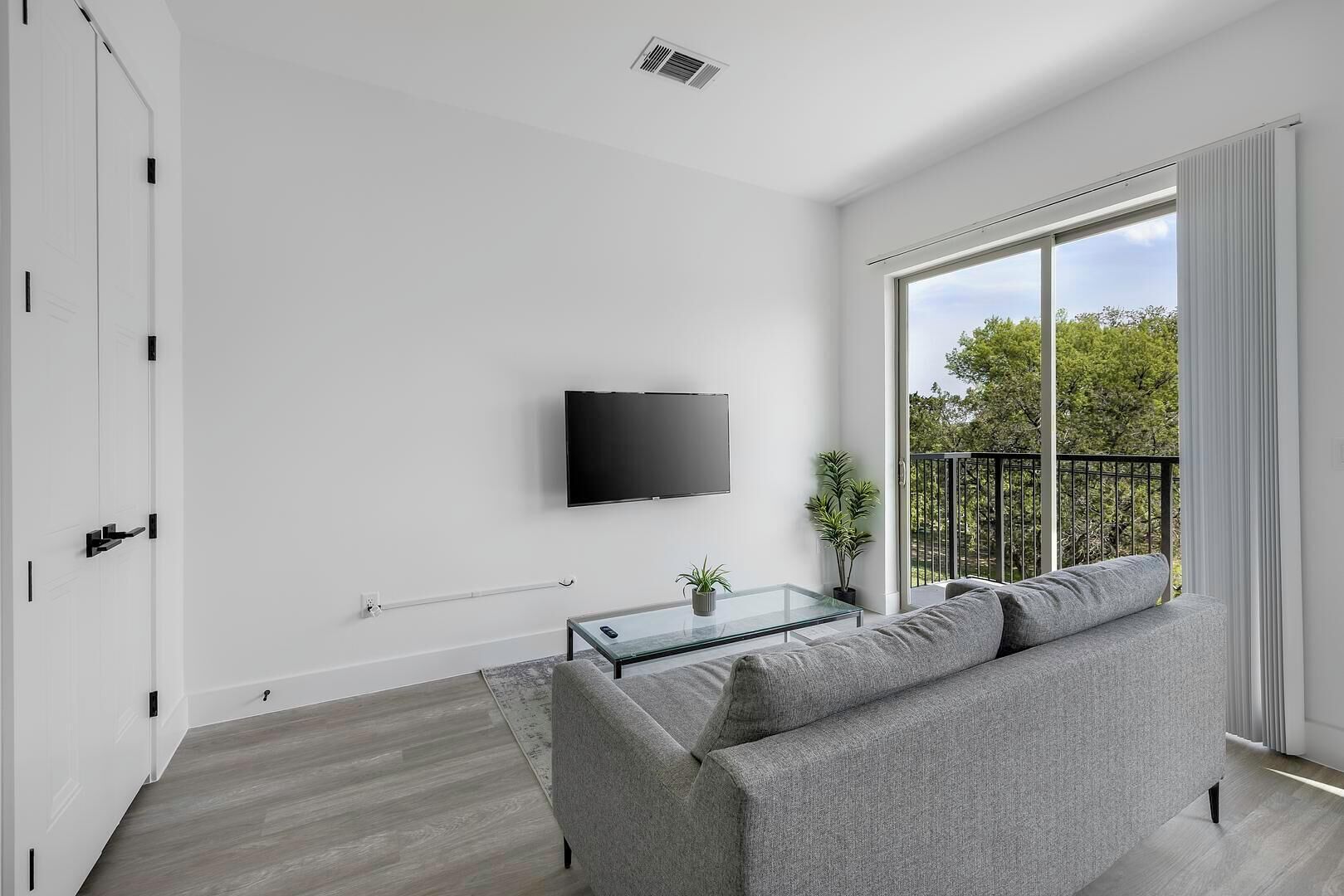 A modern living room with a grey sofa, a glass coffee table, a flat-screen TV mounted on the wall, and a balcony with a view of greenery.