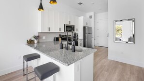 A modern kitchen with white cabinets, a granite countertop, stainless steel appliances, and black pendant lights. - Landing | Stunning Studio, Pool, Clubhouse (Denton)
