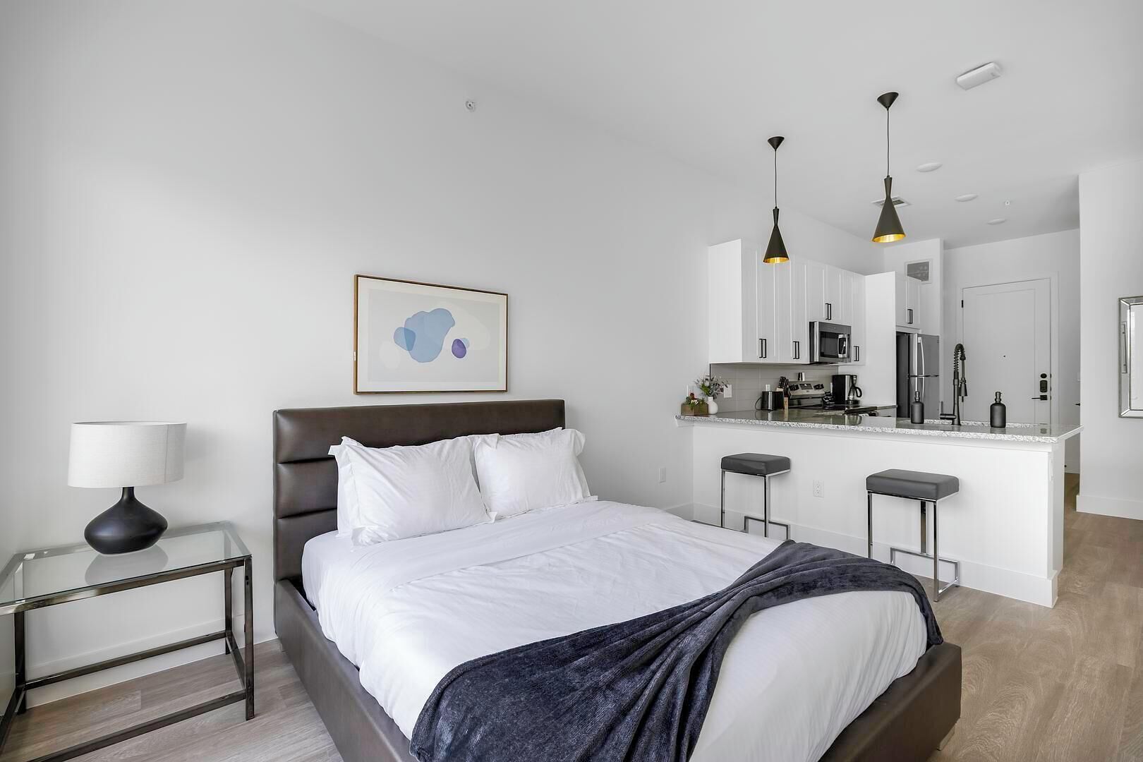 Et moderne hotelværelse med seng, sengebord, lampe og et tekøkken med mikroovn og køleskab.