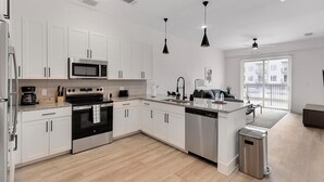A modern kitchen with white cabinets, a microwave, stove, and dishwasher. There is a small island with a sink and a trash can. The room has a large window and a ceiling fan.