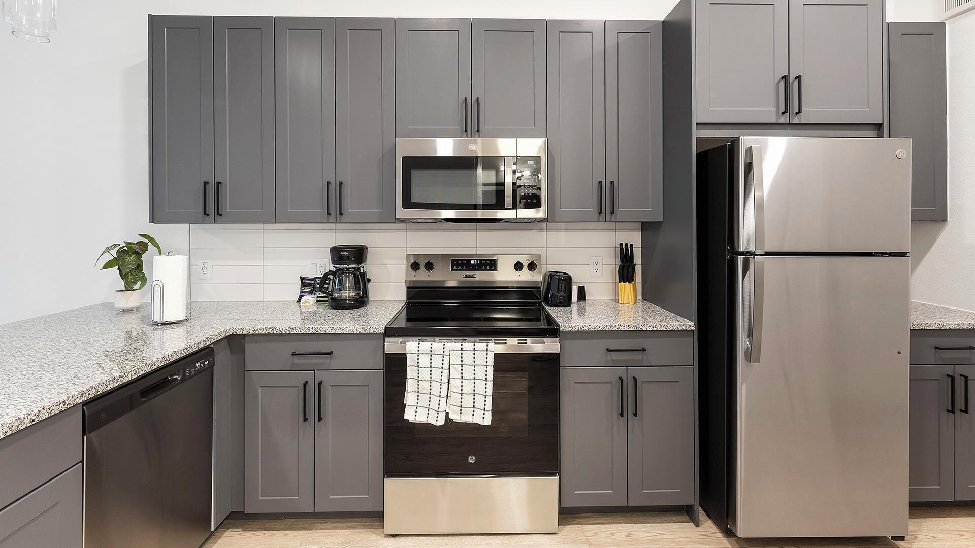 A modern kitchen with grey cabinets, a stainless steel refrigerator, a microwave, an oven, and a coffee maker.