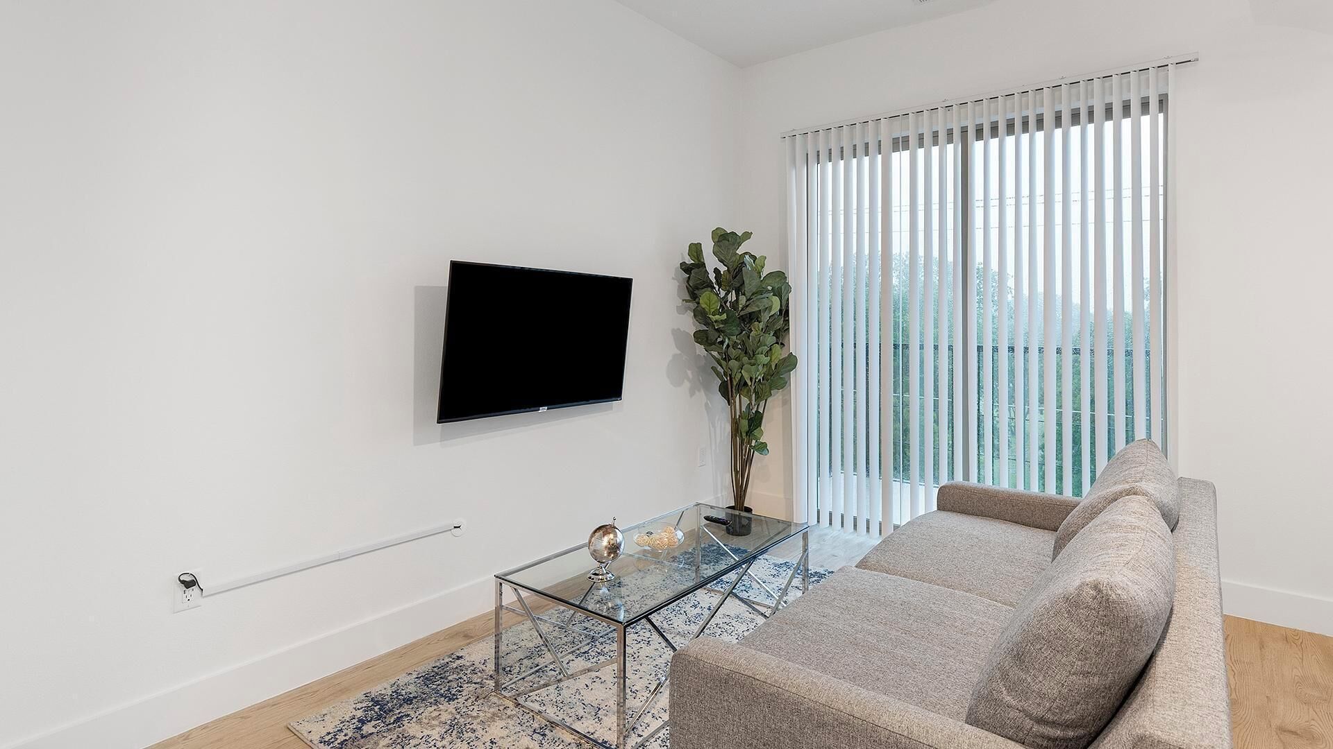 A modern living room with a flat-screen TV, a glass coffee table, a sofa, and a large window with blinds.