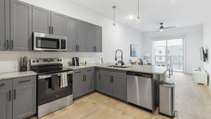 A modern kitchen with grey cabinets, stainless steel appliances, a built-in oven, microwave, and dishwasher. There is a window providing natural light and a ceiling fan. - Landing | Spectacular 1BD, Courtyard, Pool (Denton)