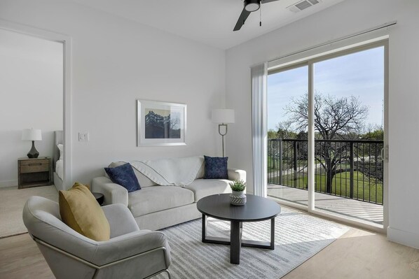 Un salon moderne avec un canapé, un fauteuil et une table basse, une grande fenêtre donnant sur un balcon et des arbres, et un ventilateur de plafond.