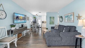 A modern living room with a grey sectional sofa, a dining area with a wooden table and chairs, and a kitchen in the background.