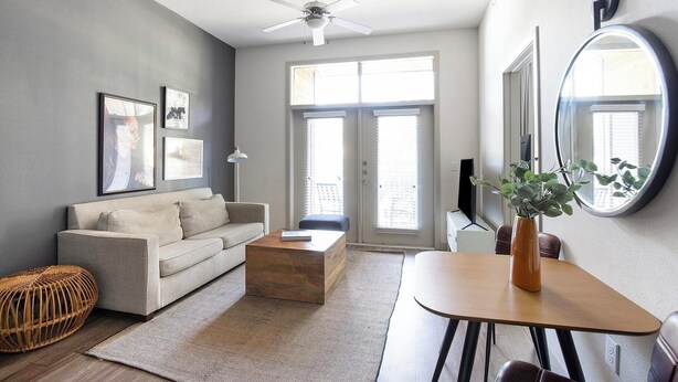 A modern living room with a grey sofa, a wooden coffee table, a dining table with a vase, and a ceiling fan. - Landing | Chic 1BD, Gym, Rooftop Lounge (San Antonio)