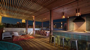 An outdoor bar area with wooden flooring, seating, and a city view at night. - Landing | Chic 1BD, Gym, Rooftop Lounge (San Antonio)