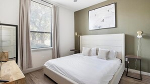 A bedroom with a bed, a window with curtains, a bedside table, a wall-mounted lamp, and a painting. - Landing | Chic 1BD, Gym, Rooftop Lounge (San Antonio)