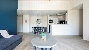 Ein modernes Wohnzimmer mit einer grauen Couch, einem runden Couchtisch und einem Essbereich mit einem schwarzen Tisch und Stühlen. Im Hintergrund ist eine Küche mit weißen Schränken und einem Edelstahl-Kühlschrank zu sehen.