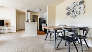 A modern kitchen with a central island, a dining area with a wooden table and black chairs, and a living room with a TV and a small side table. - Landing | Incredible 2BD, Game Room, Gym (Longboat Key)