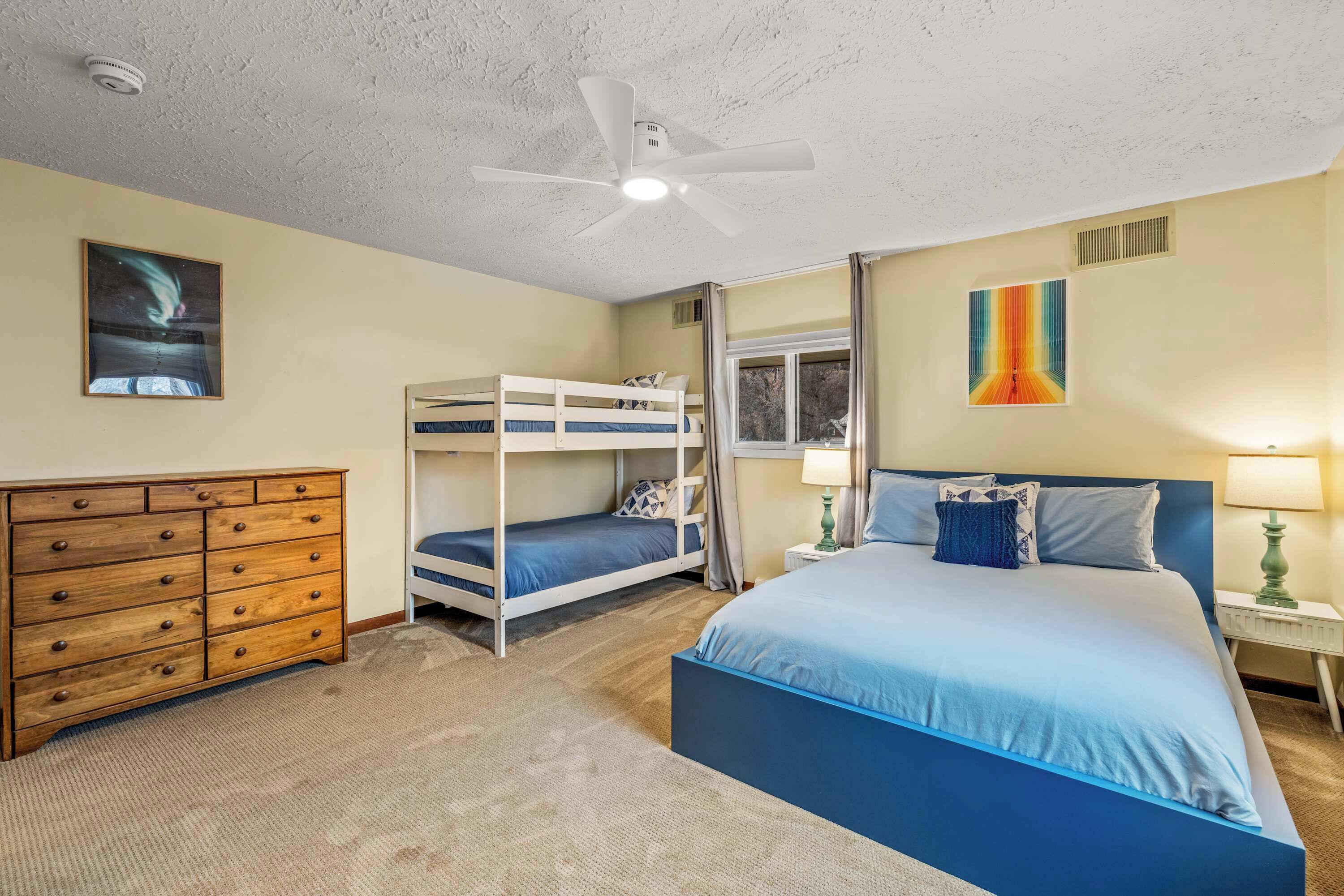 A bedroom with bunk beds, a wooden dresser, a ceiling fan, and a window with curtains.