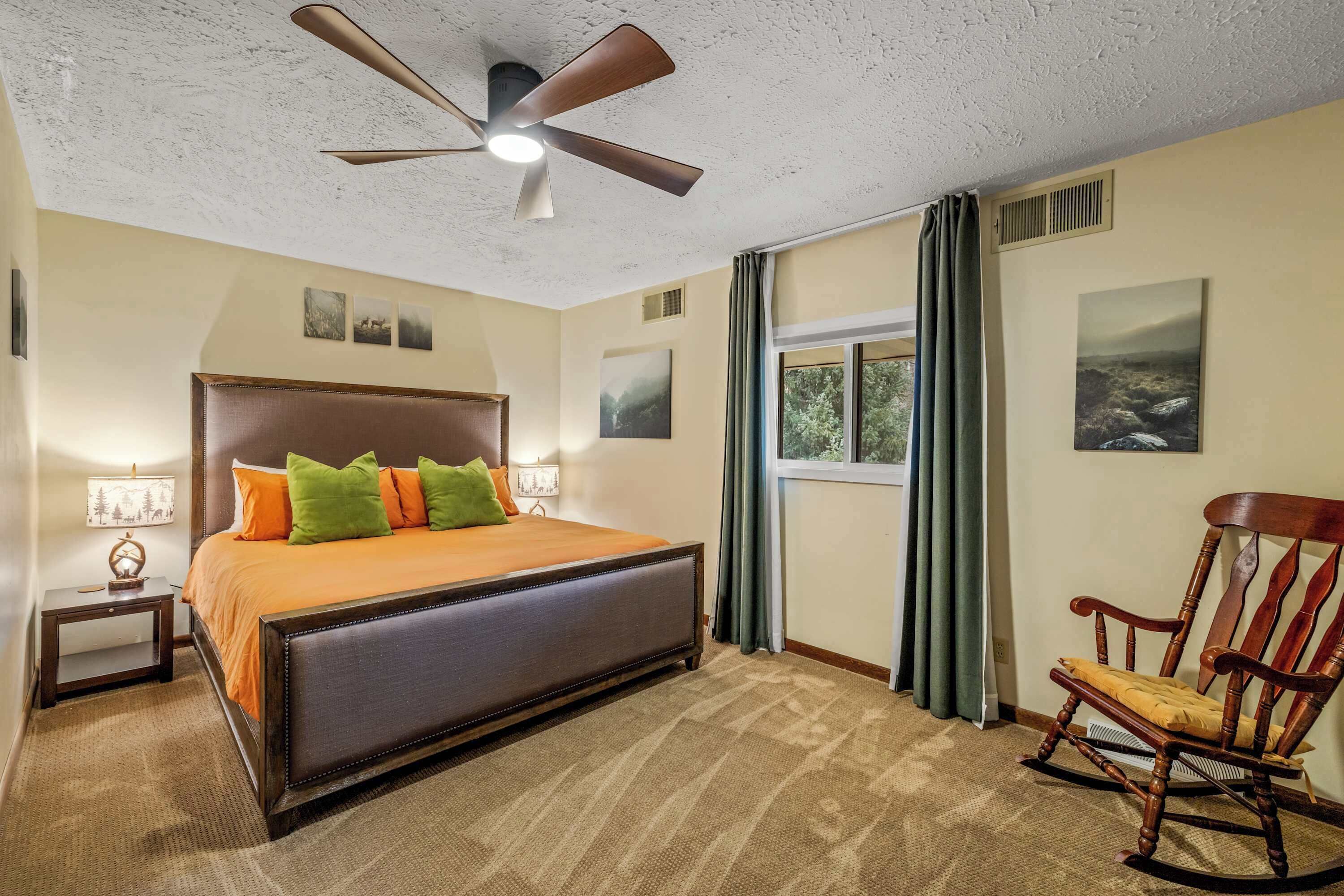 A bedroom with a large bed, a wooden rocking chair, a ceiling fan, and a window with curtains.