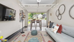Ein modernes Wohnzimmer mit einem grauen Sofa, zwei Korbstühlen, einem gemusterten Teppich und Blick auf Häuser durch eine offene Garage.
