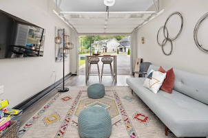 A modern living room with a grey sofa, two wicker chairs, a patterned rug, and a view of houses through an open garage.