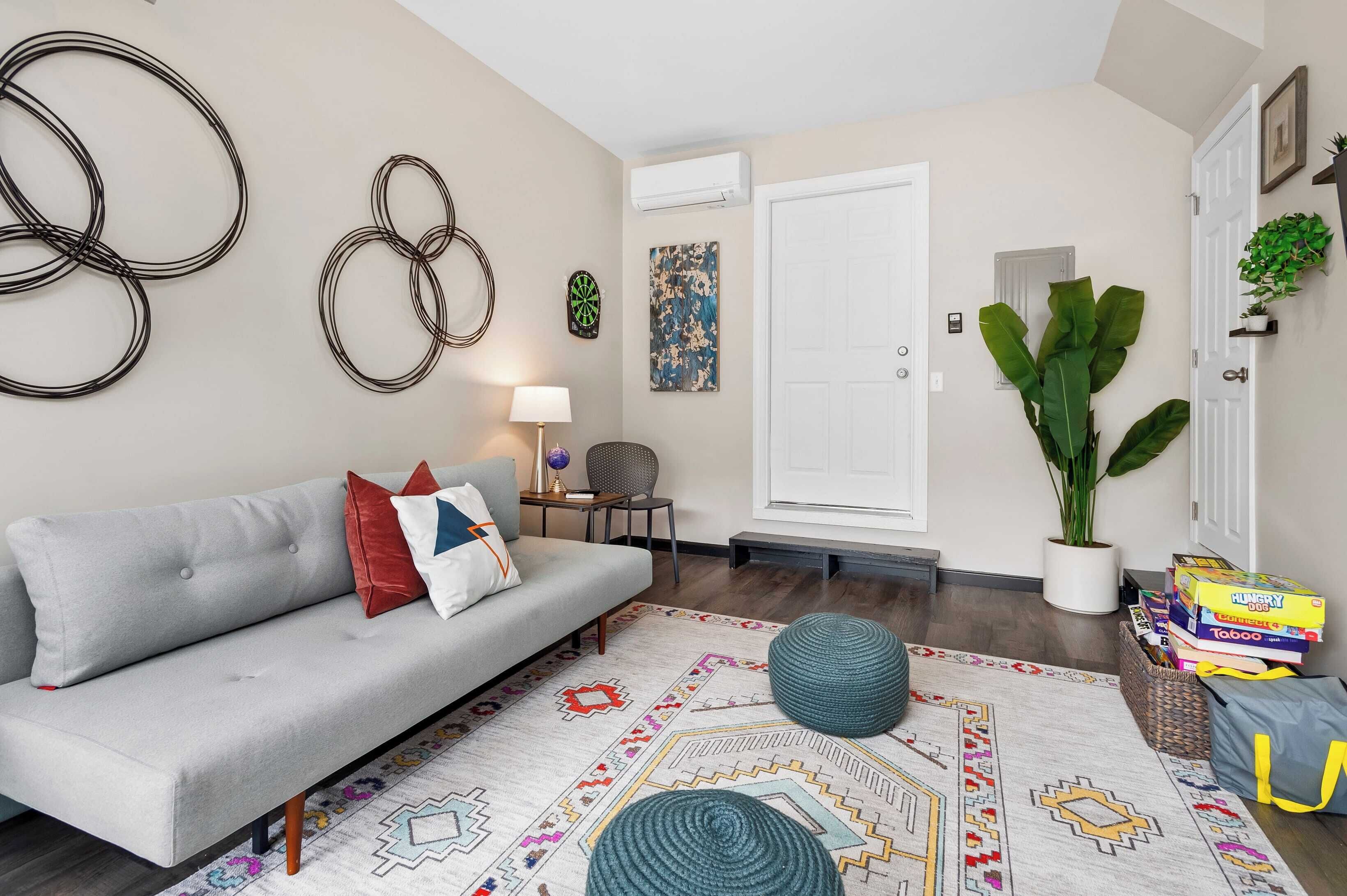 A living room with a grey sofa, a patterned rug, a potted plant, and a stack of board games.