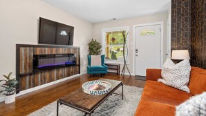 Ein Wohnzimmer mit einem an einer Holzwand montierten Flachbildfernseher, einem blauen Sessel, einem orangefarbenen Sofa und einem Couchtisch mit dekorativer Ablage.
