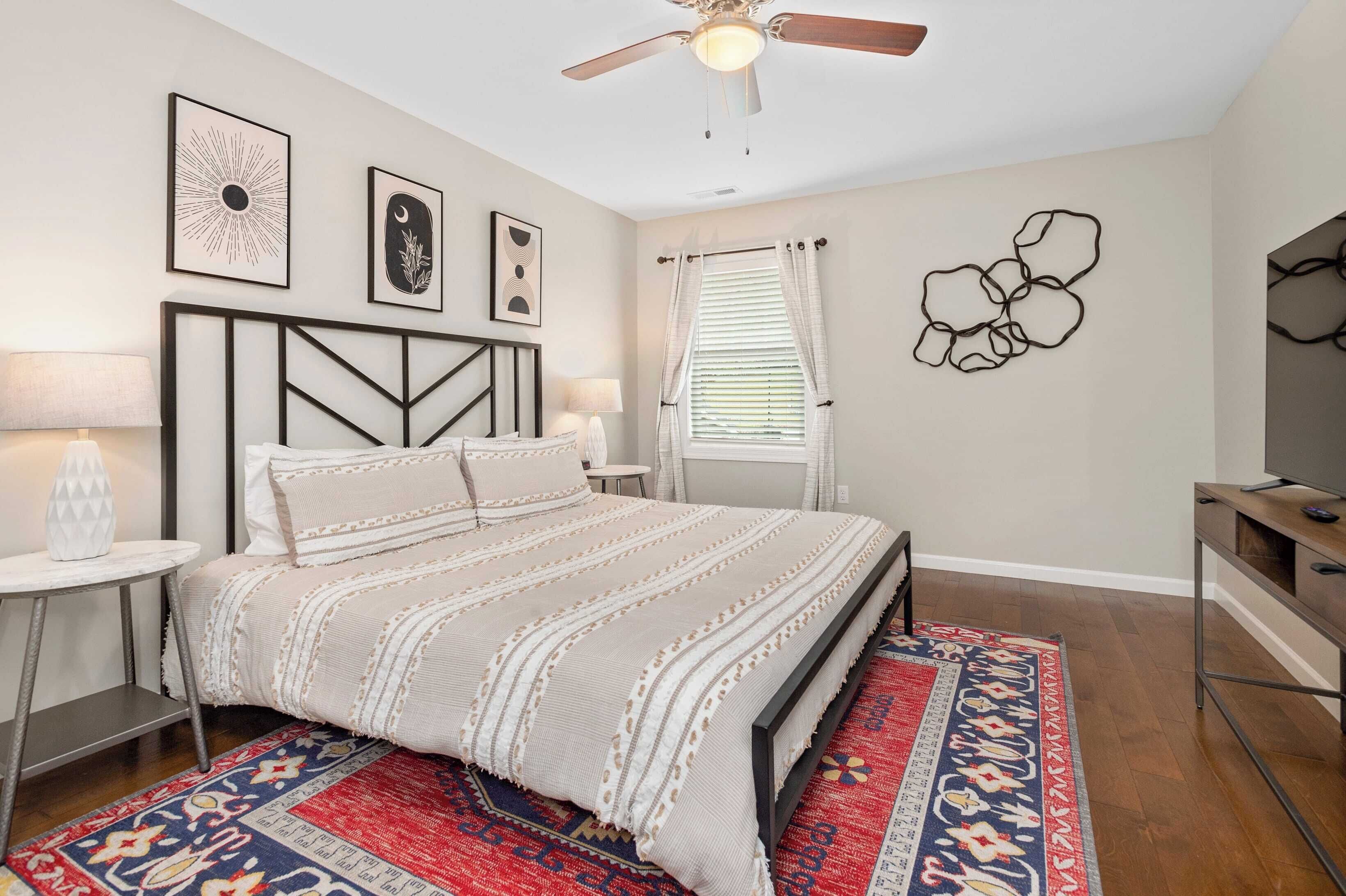 A bedroom with a bed, bedside tables, a ceiling fan, a window with curtains, and wall art.