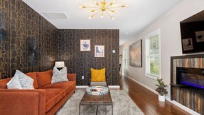 Una sala de estar con sofá, mesa de centro y cojines decorativos. Hay cuadros enmarcados en la pared, una lámpara en el techo y un televisor sobre un mueble de madera.