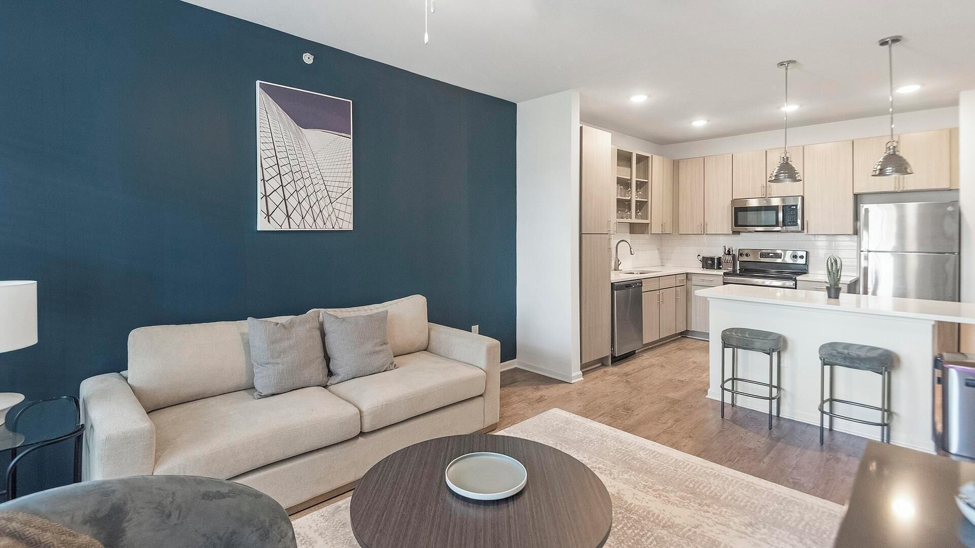 A modern living room with a sofa, a coffee table, and a kitchen area with a microwave, refrigerator, and bar stools.
