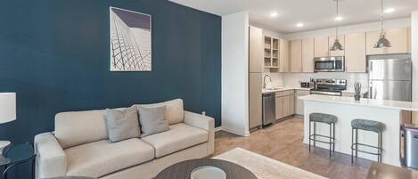 A modern living room with a sofa, a coffee table, and a kitchen area with a microwave, refrigerator, and bar stools.