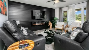 A modern living room with leather sofas, a fireplace, a mounted TV, and a ceiling fan. - Roadrunner Ridge | 3/3, Game Room, Hot Tub, Fire Pit & Grill, Pets w/ fee (Fredericksburg)