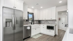 A modern kitchen with white cabinets, stainless steel appliances, a water dispenser, and a gray tiled backsplash. - Roadrunner Ridge | 3/3, Game Room, Hot Tub, Fire Pit & Grill, Pets w/ fee (Fredericksburg)