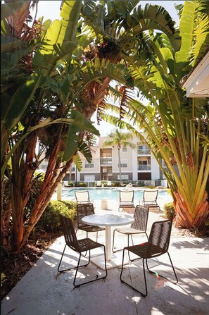An outdoor seating area with a round table and four chairs, surrounded by large banana trees and a pool in the background.