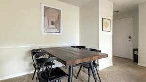 Un coin repas avec une table en bois et des chaises noires, un tableau encadré au mur et une porte à l’arrière-plan.