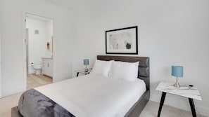 Ein Hotelzimmer mit Bett, Nachttisch mit Lampe, einem gerahmten Bild an der Wand und einem durch eine offene Tür einsehbaren Badezimmer.