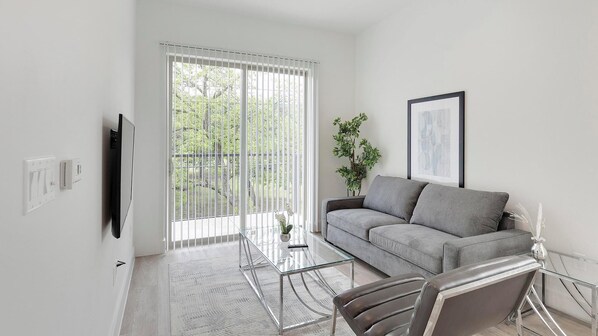 Ein modernes Wohnzimmer mit grauem Sofa, einem Glastisch und einem Ledersessel. Ein großes Fenster mit Jalousien, ein Topfpflanze und ein gerahmtes Bild sind ebenfalls zu sehen.