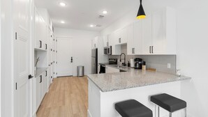 A modern kitchen with white cabinets, granite countertops, a stainless steel refrigerator, a microwave, and a coffee maker. There are two black bar stools and a hanging pendant light. - Landing | Spectacular 1BD, Pool, Clubhouse (Denton)