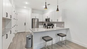 A modern kitchen with white cabinets, a stainless steel refrigerator, a microwave, and a granite countertop. There are two bar stools and a trash can. - Landing | Stylish 1BD, Pool, Gym (Denton)