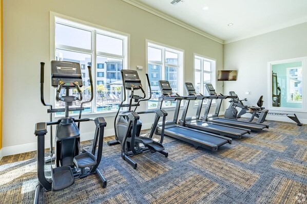 A gym with treadmills, elliptical machines, and a stationary bike, large windows, and a carpeted floor.