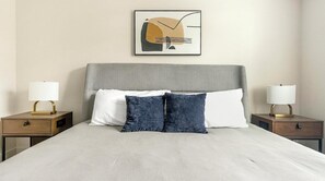 Un llit ben arreglat amb un panell frontal gris, dues taules de nit amb làmpades i una obra d'art abstracta emmarcada sobre el llit.