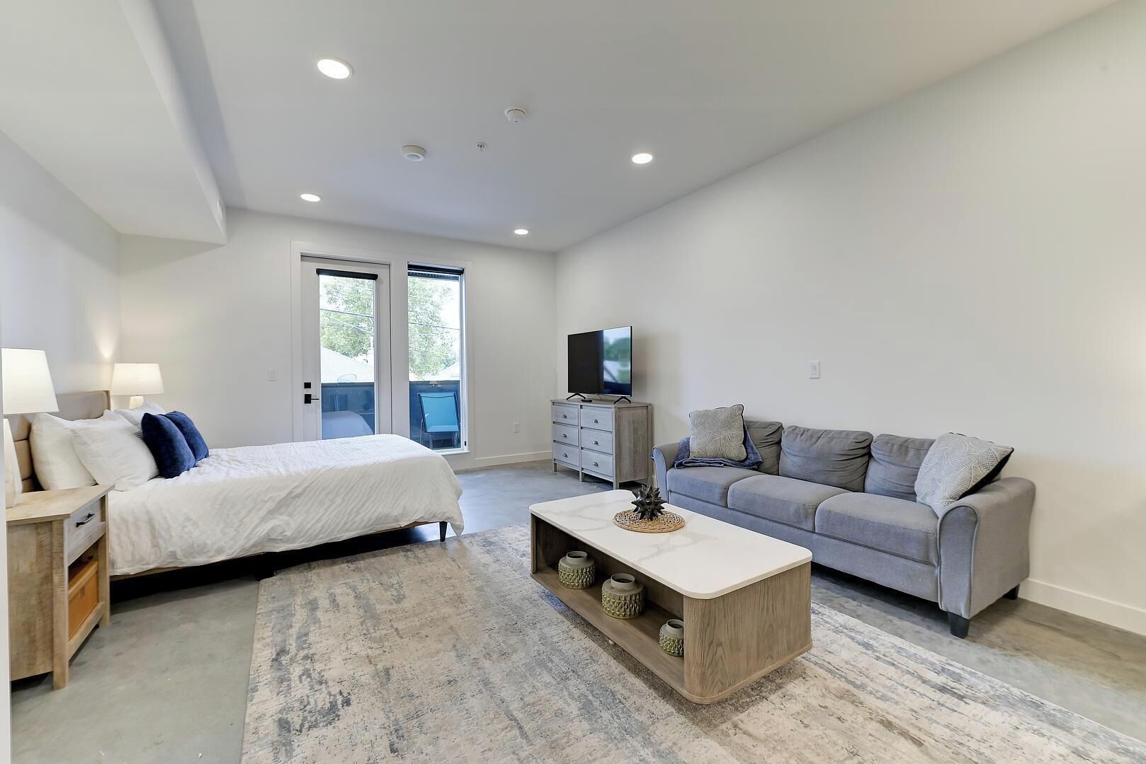 A modern bedroom with a bed, a grey sofa, a TV, and a large window.
