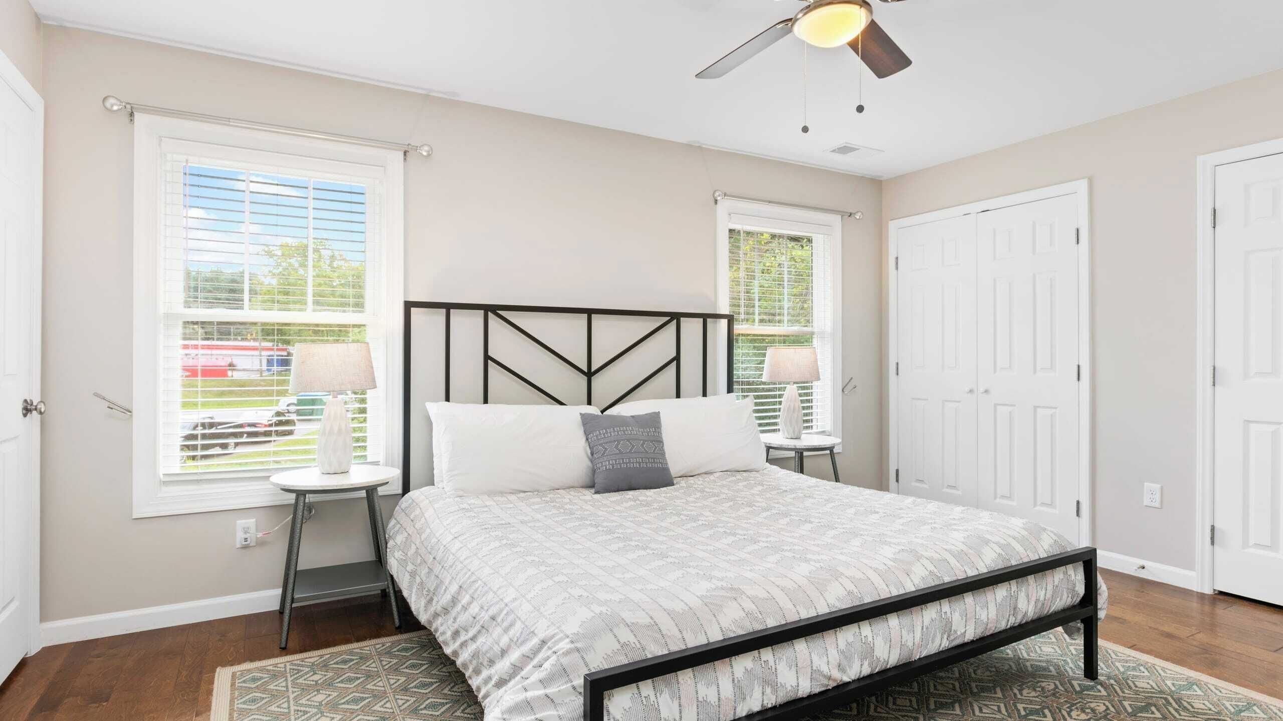 A bedroom with a bed, two bedside tables, a ceiling fan, and two windows with blinds.
