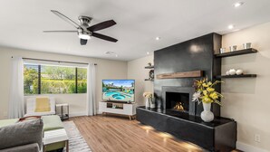 A modern living room with a fireplace, a ceiling fan, and a large window with blinds.