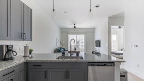 A modern kitchen with grey cabinets, a granite countertop, a coffee maker, and a dishwasher. - Landing | Modern 1BD, Courtyard, Pool (Denton)