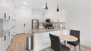 A modern kitchen with white cabinetry, stainless steel appliances, and a granite countertop. There is a dining area with a round table and two chairs. - Landing | Chic 2BD, Gym, Courtyard (Denton)