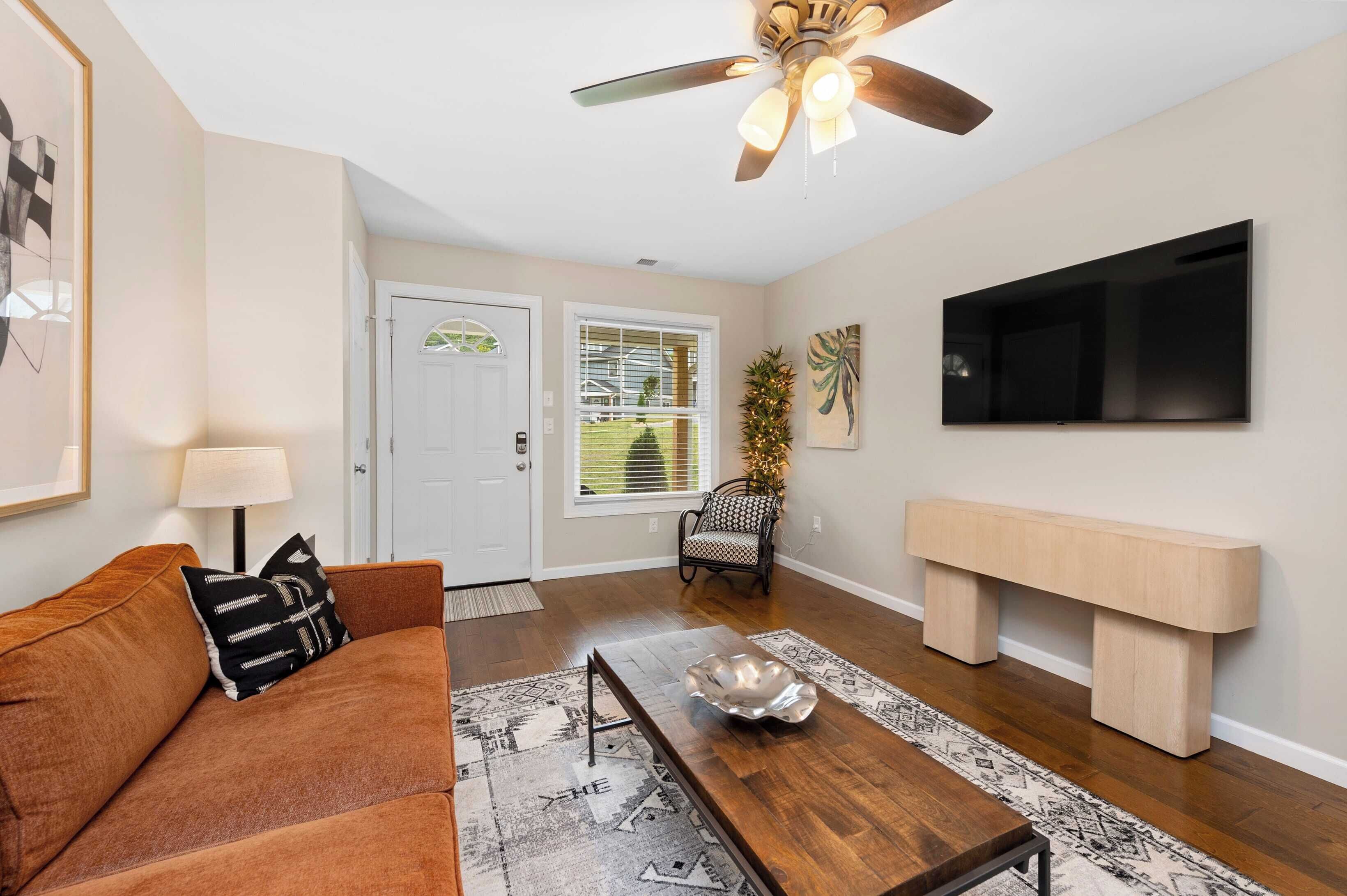 A living room with a brown sofa, a coffee table, a TV mounted on the wall, a ceiling fan, and a door with a window.