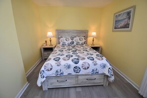 En seng med blomstret sengetæppe, to sengeborde med lamper og et billede i ramme på væggen.