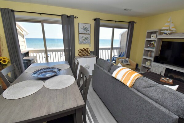 A living room with a sofa, a dining table with placemats, a TV, and a view of the sea.